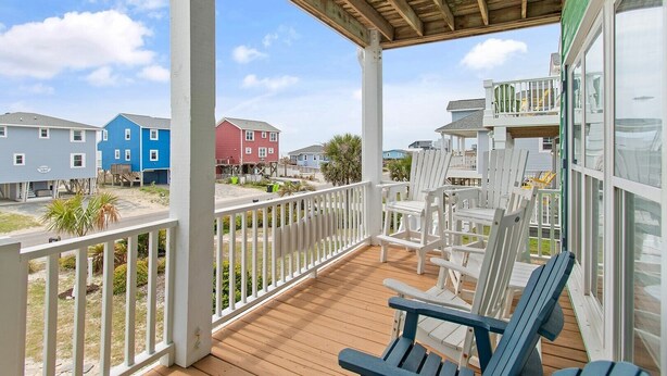 A covered porch with white railings and wooden flooring, overlooking a residential area with colorful houses. - Oceanview Key Rest on Oak Island: Spacious 6BR, 5BA home for 15 guests with pool. (Oak Island)