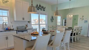 A kitchen with white cabinets, a granite countertop, and a dining area with white chairs. - Oceanview Key Rest on Oak Island: Spacious 6BR, 5BA home for 15 guests with pool. (Oak Island)