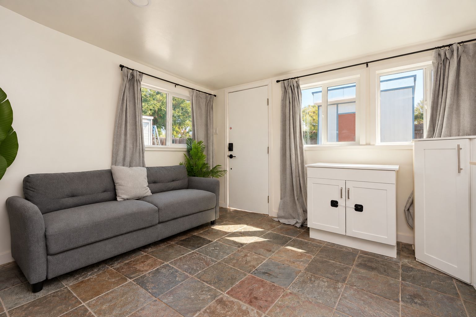 Ein Zimmer mit einer grauen Couch, einem weißen Schrank und einem großen Fenster mit Vorhängen.
