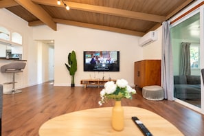 Ein modernes Wohnzimmer mit Holzboden, einem an der Wand befestigten Fernseher, einem Esstisch mit Blumenvase und einem Sofa.