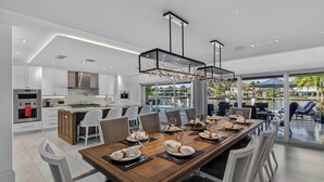 Cozinha moderna com armários brancos, uma ilha central e uma área de jantar com uma mesa de madeira comprida para seis pessoas.