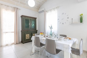 Zona pranzo con tavolo apparecchiato per quattro, un armadietto verde, una sveglia a parete e una finestra con tende.