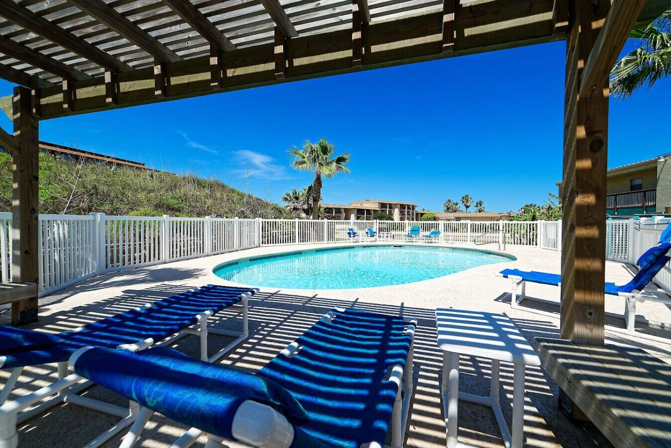 A pool area with lounge chairs, a wooden pergola, and a view of a building and palm trees.