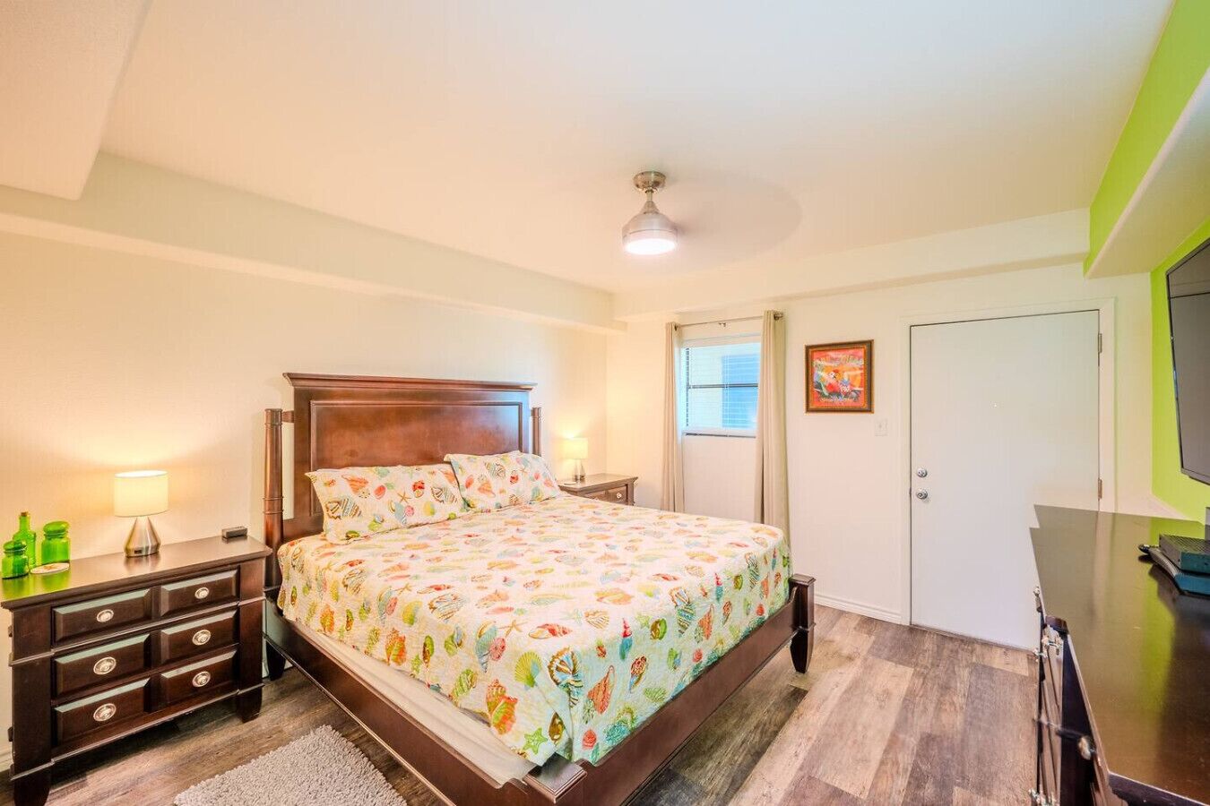 Un dormitorio con una cama de madera, una mesita de noche con lámpara, un aparador y una puerta con un cuadro.