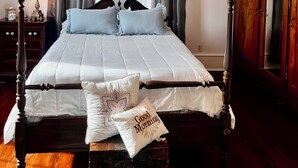 A neatly made bed with a white and gray bedspread, two pillows, and a 'Good Morning' pillow. A chandelier hangs above the bed, and a wooden cabinet is to the right.