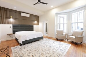 Une chambre à coucher comprenant un lit, un ventilateur de plafond, une grande fenêtre et un coin salon avec deux fauteuils.