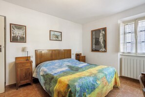 Un dormitorio con cama, mesita de noche, lámpara, ventana y dos cuadros en la pared.