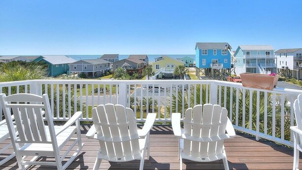 A deck with white Adirondack chairs, a view of beach houses, and a clear blue sky. - Experience ocean views in this spacious 4-bed, 3-bath home on Oak Island. (Oak Island)