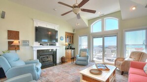 A living room with a fireplace, television, and large windows overlooking a balcony. - Experience ocean views in this spacious 4-bed, 3-bath home on Oak Island. (Oak Island)
