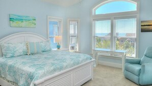 A bedroom with a bed, a chair, a ceiling fan, and a large window with blinds. - Experience ocean views in this spacious 4-bed, 3-bath home on Oak Island. (Oak Island)