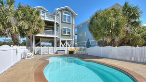 A swimming pool with a white fence, a two-story building with balconies, and palm trees. - Experience ocean views in this spacious 4-bed, 3-bath home on Oak Island. (Oak Island)