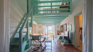 Eine Treppe mit türkisfarbenen Geländern führt zu einem Balkon, einem Essbereich mit Tisch und Stühlen sowie einem Wohnbereich mit Sofa und Fernseher.