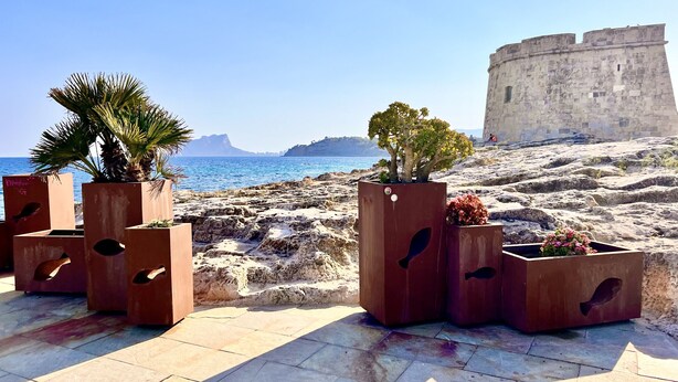 Outdoor planters with plants, a stone pathway, and a historic stone building with a view of the sea. - Boho Luxury Apartment · Moraira Centre THE MORAIRA COLLECTION - 2 (Moraira)