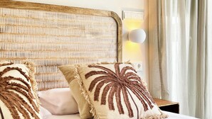 A bed with a woven headboard, two pillows with fringe details, a bedside table with a lamp, and a window with curtains. - Boho Luxury Apartment · Moraira Centre THE MORAIRA COLLECTION - 2 (Moraira)