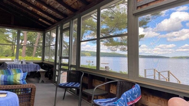 Et overdækket udendørs opholdsområde med borde og stole, store vinduer med udsigt til et søområde, og et træloft.