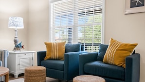 A living room with a blue sofa set, two matching cushions, a white side table with a lamp, and a woven ottoman. - Experience Oak Island at Stitch by the Sea, a cozy 3-bedroom retreat for 10. (Oak Island)