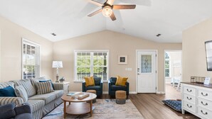 A spacious living room with a high ceiling, a ceiling fan, a sofa, a coffee table, a TV, and a door leading to another room. - Experience Oak Island at Stitch by the Sea, a cozy 3-bedroom retreat for 10. (Oak Island)