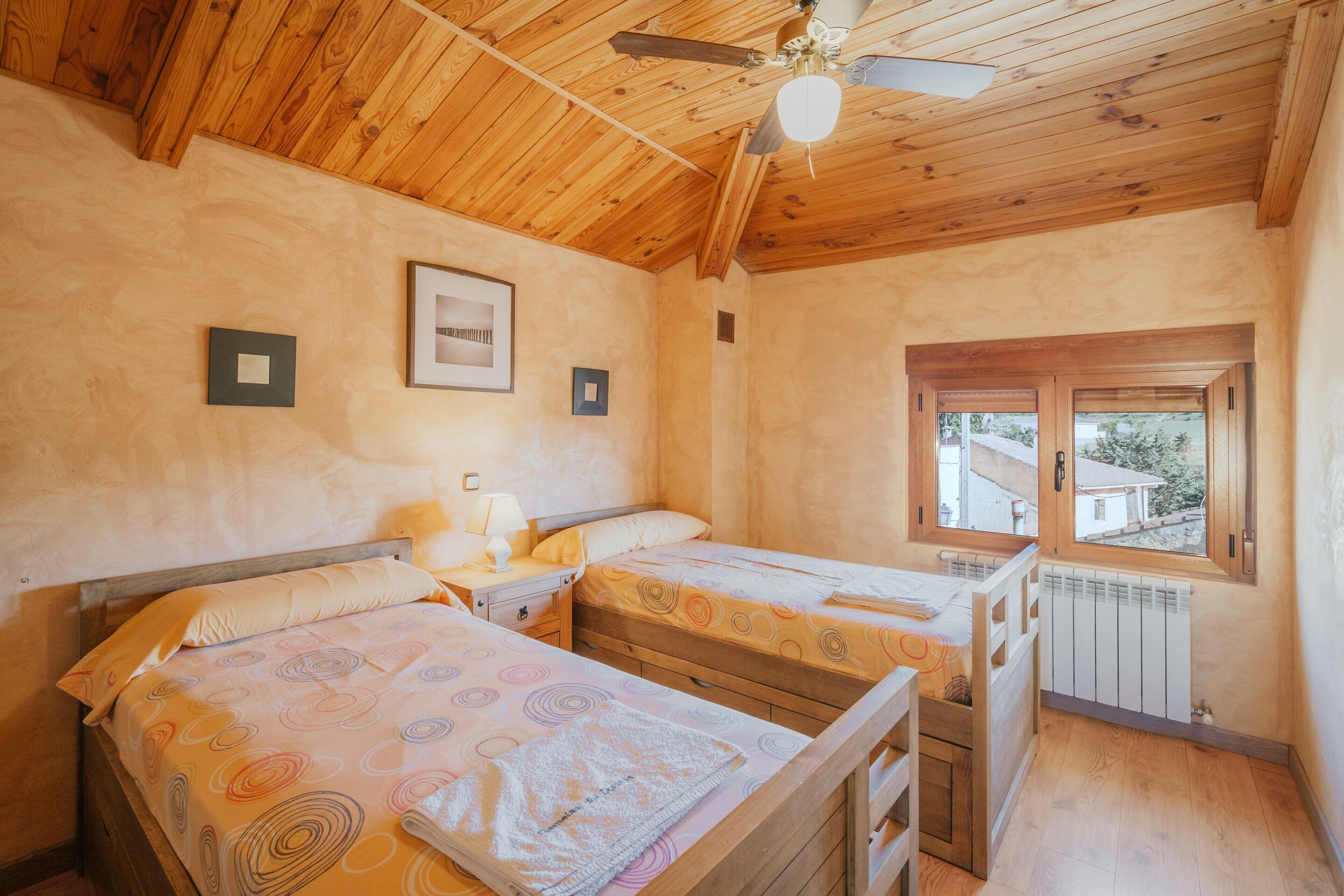 Ein Zimmer mit Holzboden und -wänden, zwei Betten mit gemusterten Bettbezügen, ein Nachttisch mit Lampe, ein Fenster mit Blick und ein Deckenventilator.