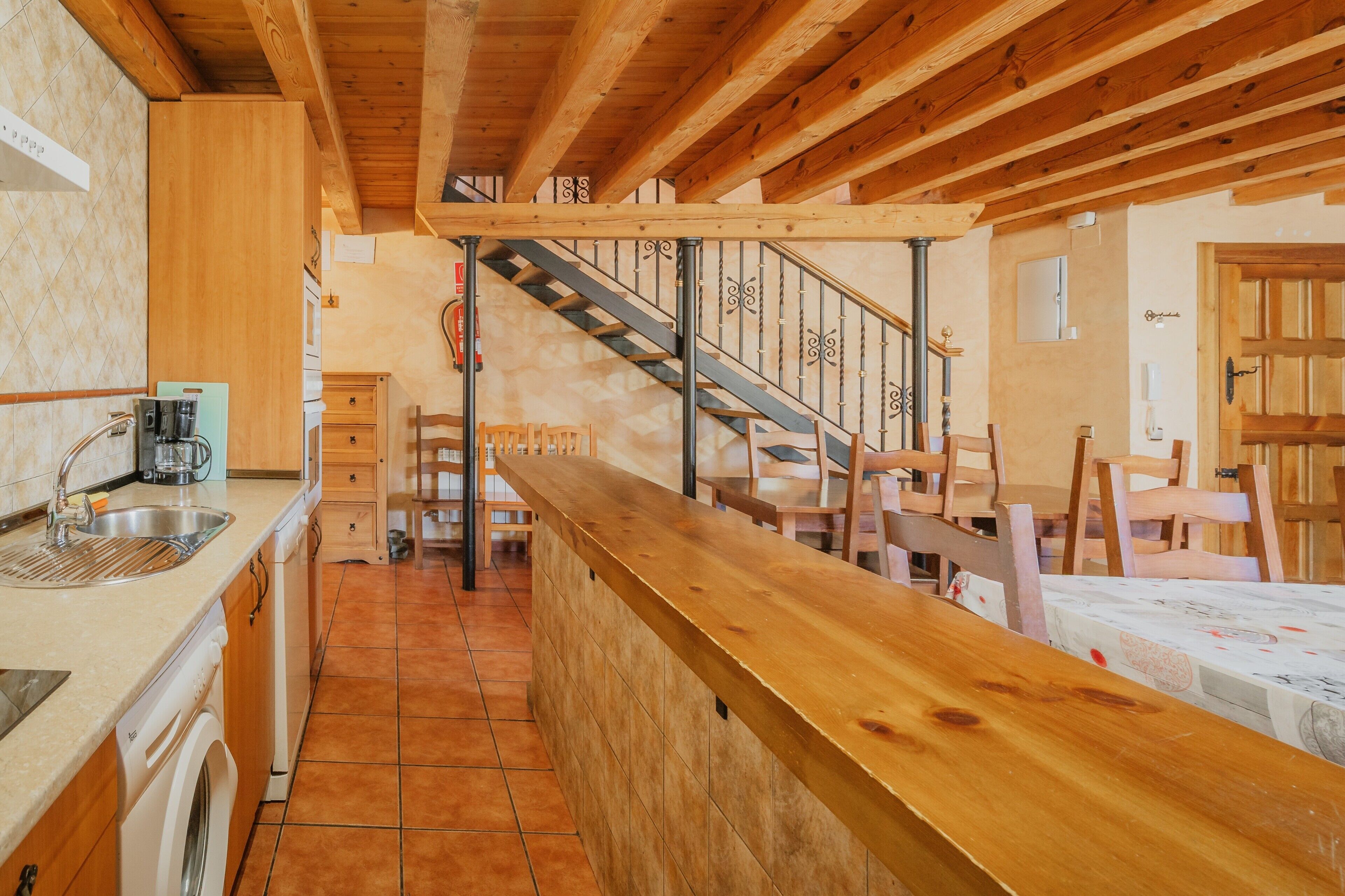 Una cocina con armarios de madera, un lavamanos y una estufa. Hay una encimera de madera con sillas y una escalera que sube a otro nivel.