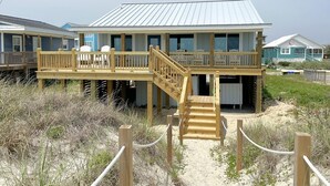 Een vakantiehuis aan het strand met een houten terras, een trap naar een overdekt gedeelte en een gebouw met een metalen dak.