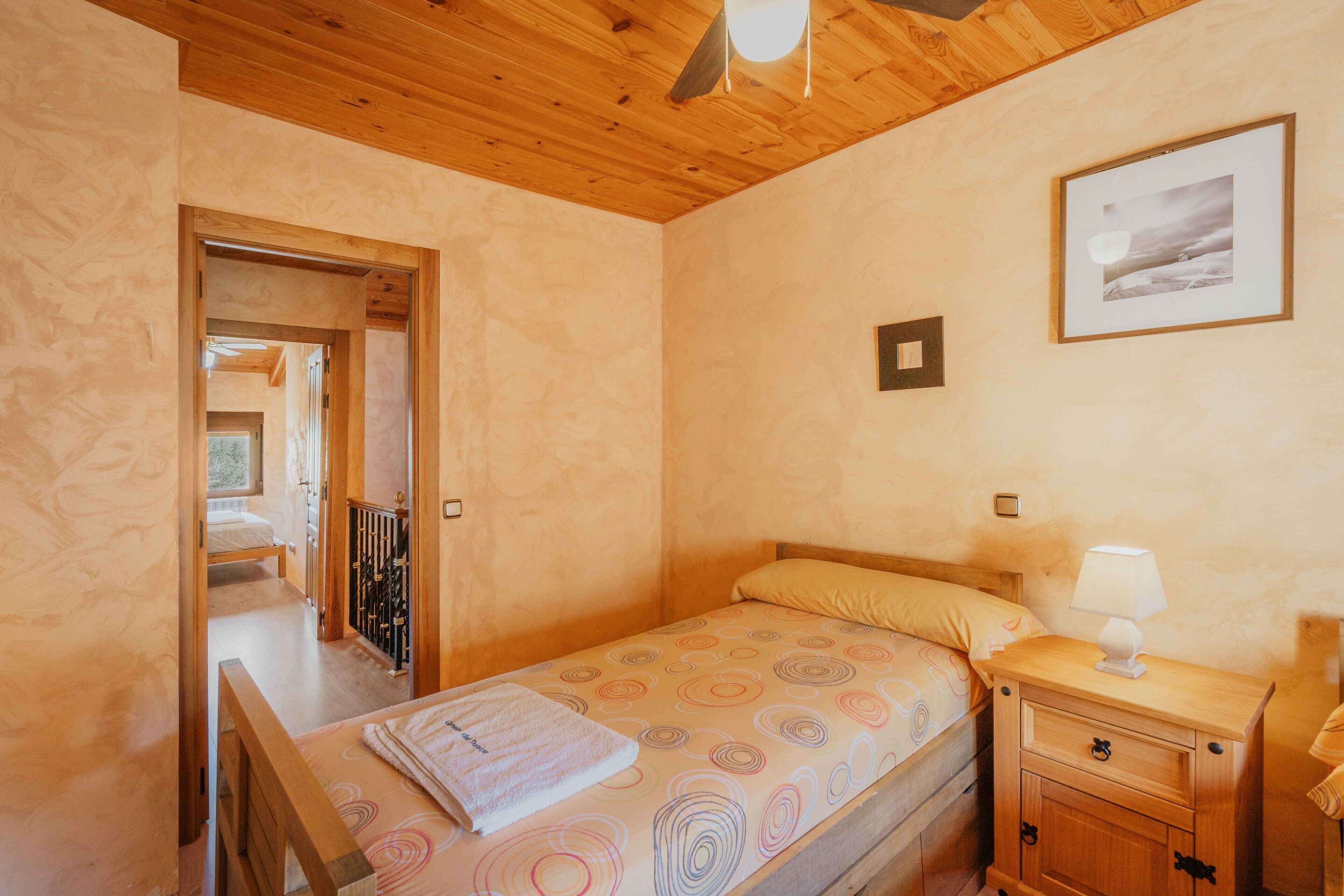 Une chambre avec un plafond en bois, un lit recouvert d’une couvre-lit à motifs, une table de chevet avec une lampe et un tableau encadré au mur.