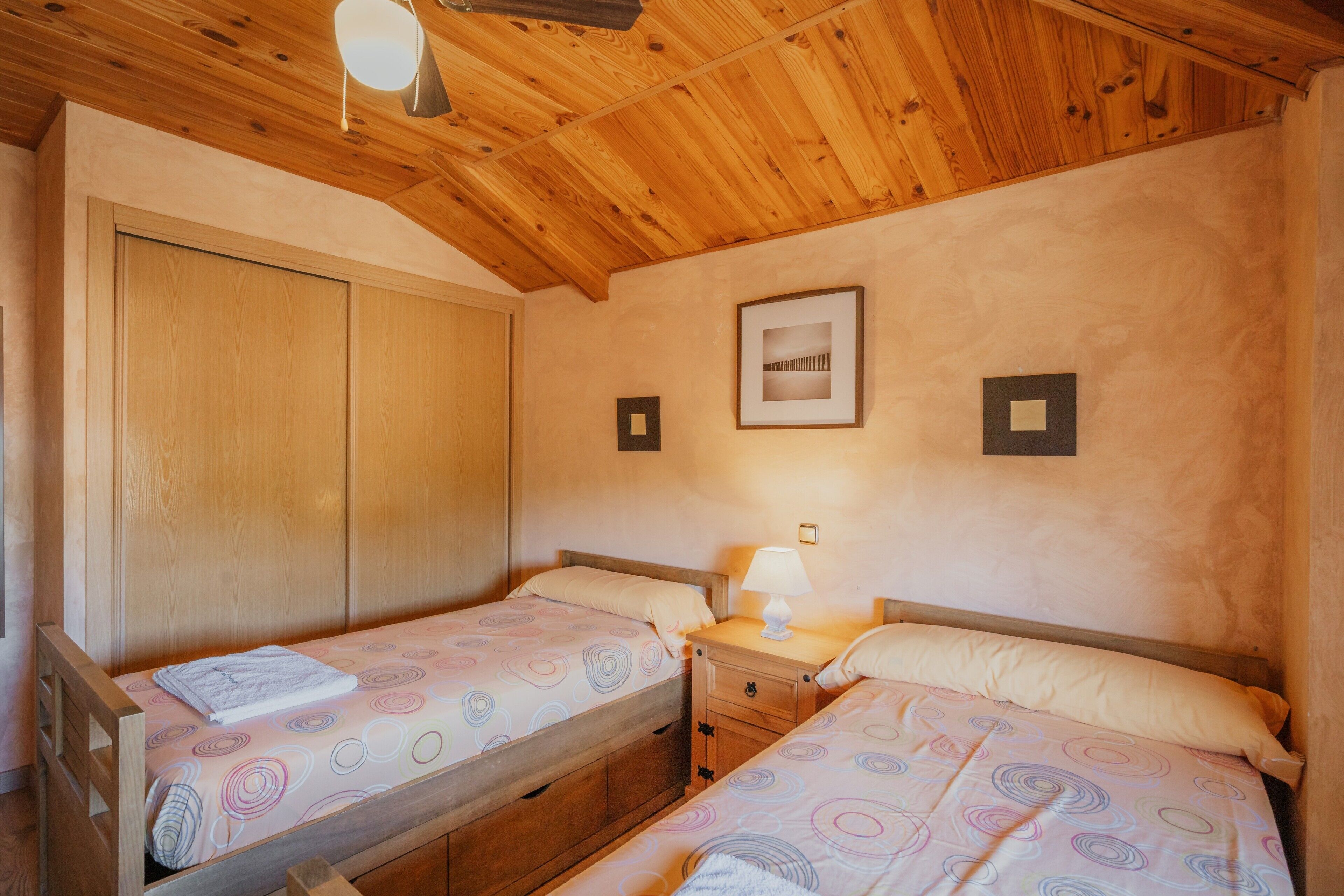 Una habitació amb dos llits de fusta, un armari de fusta, una tauleta de nit amb una làmpada i un quadre al mur.