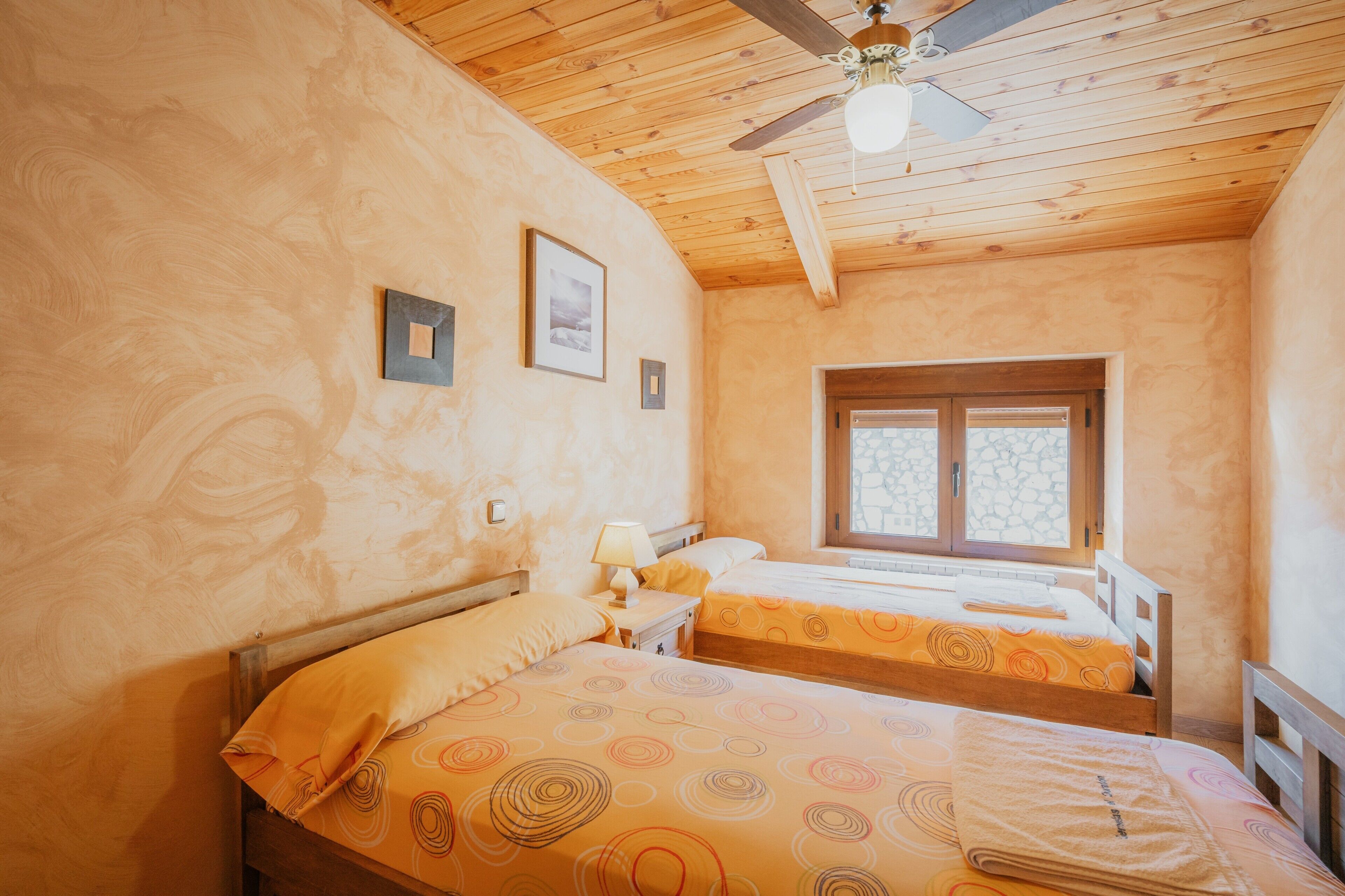 Una habitación con dos camas, techo de madera, una ventana y un ventilador de techo.