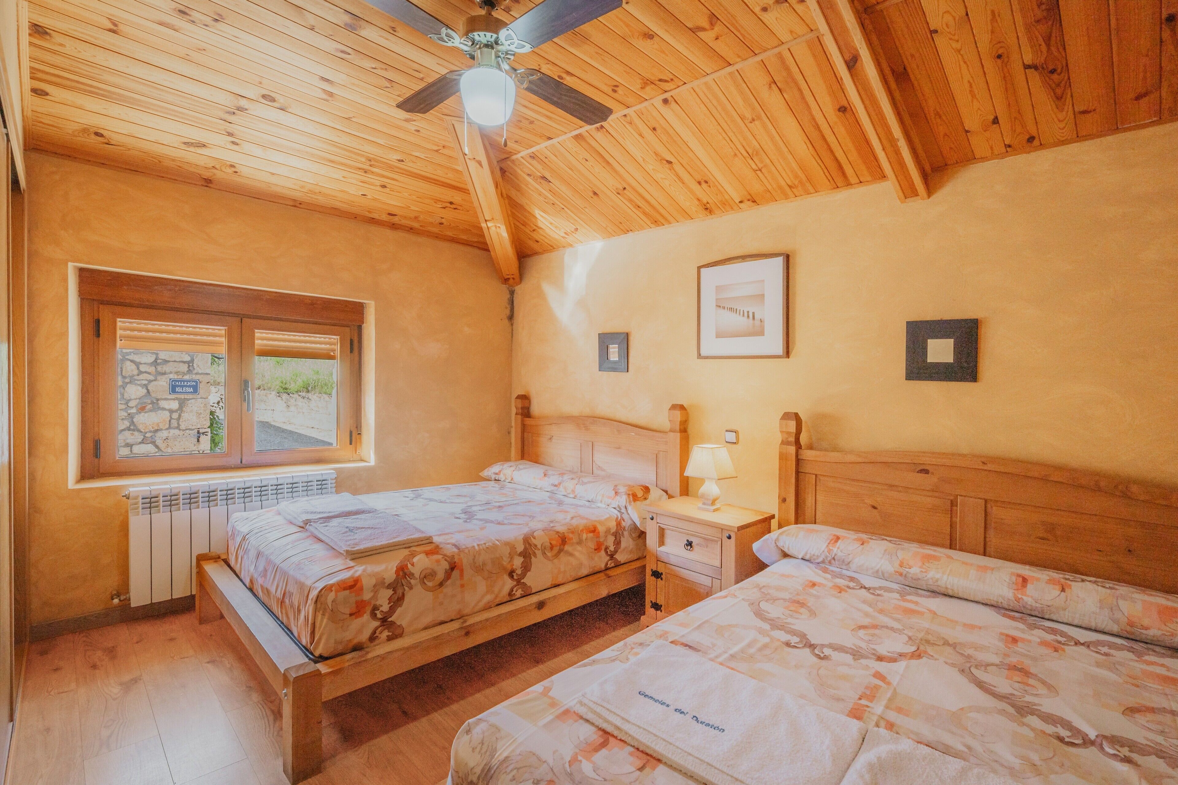 Un dormitorio con dos camas, un ventilador de techo, una ventana con marco de piedra y un cuadro en la pared.