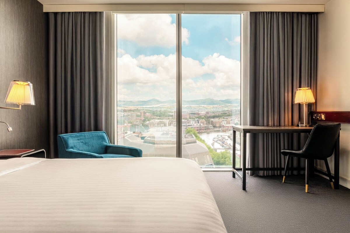 Hotelkamer met een bed, een bureau, twee stoelen, een lamp en een groot raam met uitzicht op de stad.