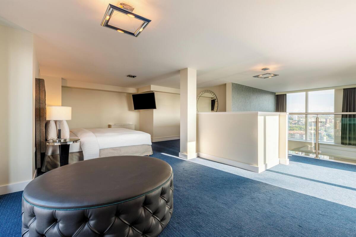 Een hotelkamer met een bed, een rond poefje, een televisie, een spiegel en een groot raam met uitzicht op de stad.
