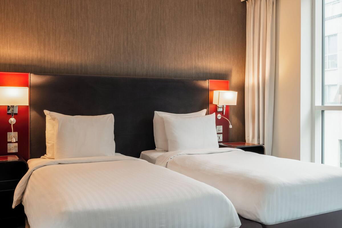 Een hotelkamer met twee bedden, een houten hoofdeinde, twee bedlampjes en een raam met gordijnen.