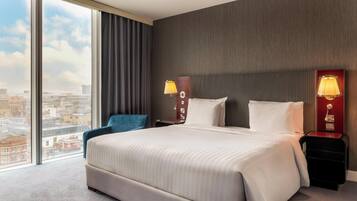 Habitación de hotel con una cama grande, dos mesitas de noche con lámparas, una silla azul y vistas a la ciudad a través de amplios ventanales.