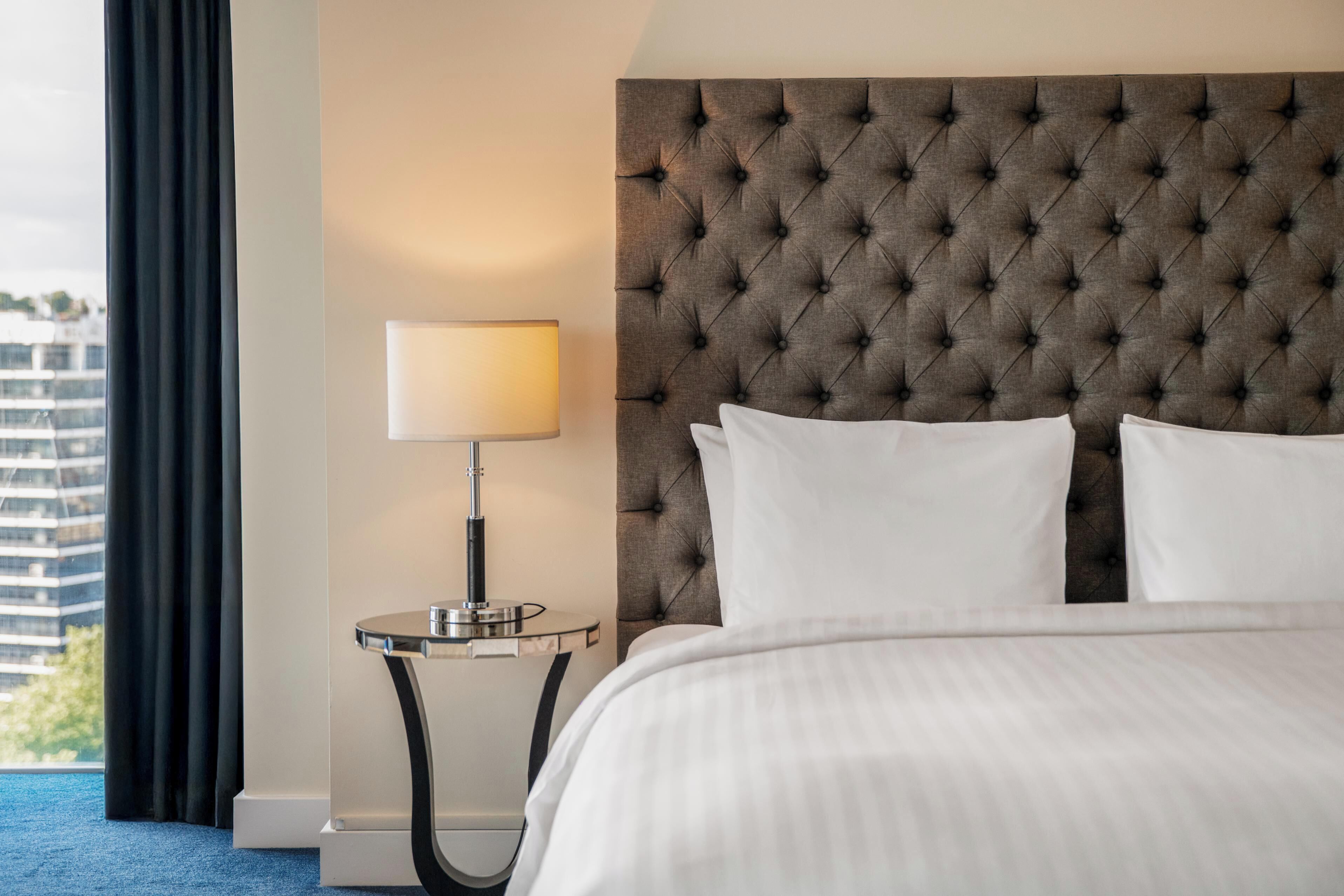 A hotel room with a tufted headboard, a bedside lamp, a bed with white linens, and a view of a building through a window.