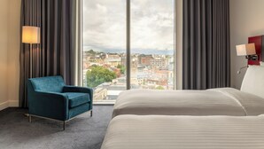 Habitación de hotel con una cama grande, un sillón azul, vista a la ciudad a través de un ventanal amplio y una mesita de noche roja.