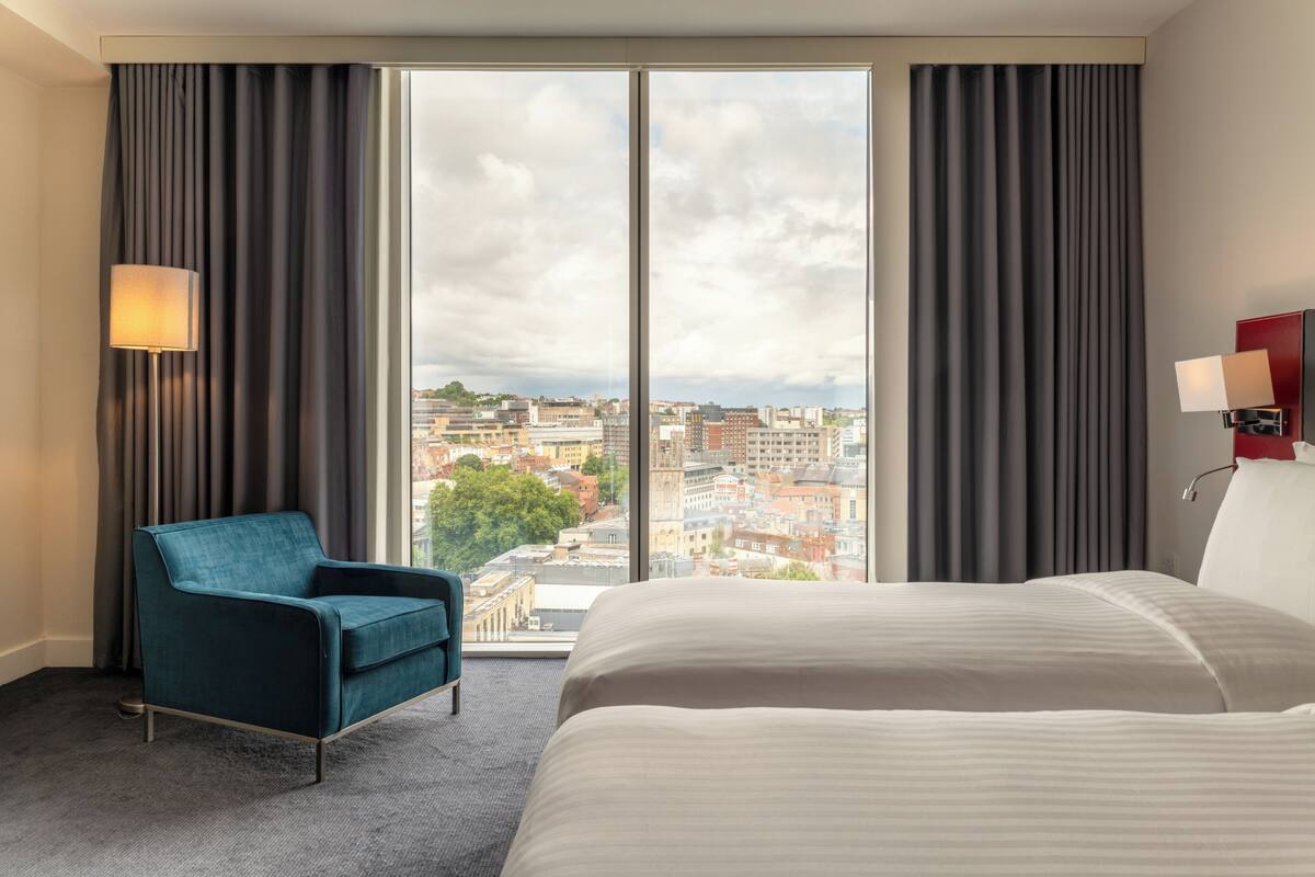 Een hotelkamer met een groot bed, een blauwe fauteuil, stadszicht door een groot raam en een rode nachtkastje.