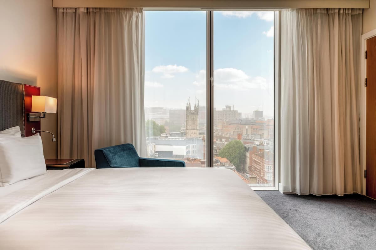 Hotelkamer met een groot bed, een stoel, een nachtkastje met een lamp en uitzicht op de stad door middel van kamerhoge ramen.