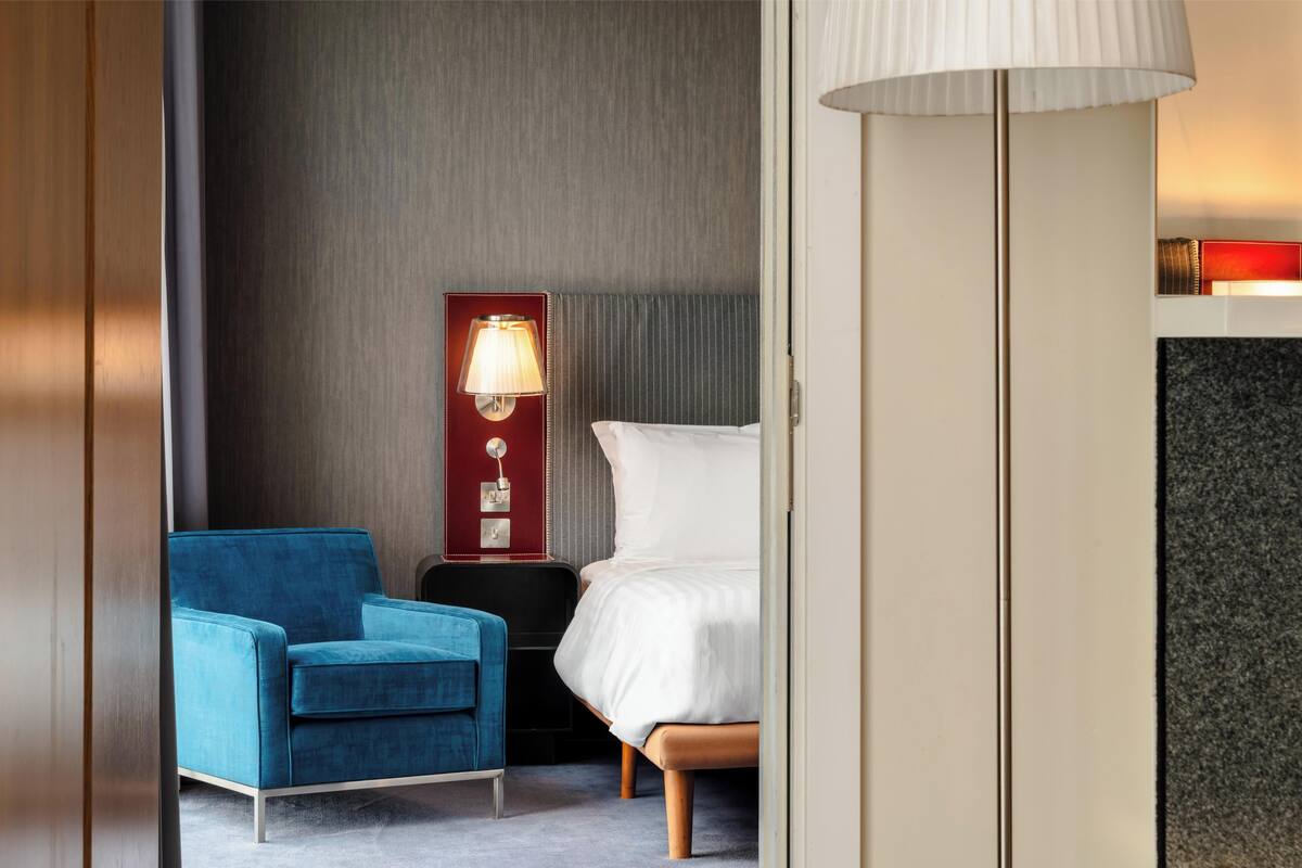 Een hotelkamer met een bed, een blauwe fauteuil, een nachtkastje met een lamp en een rode muur.