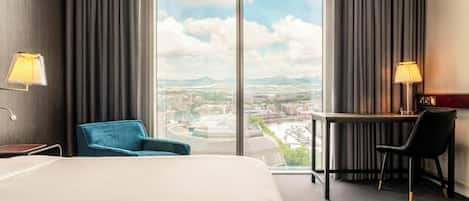 A hotel room with a bed, a desk, two chairs, a lamp, and a large window with a city view.