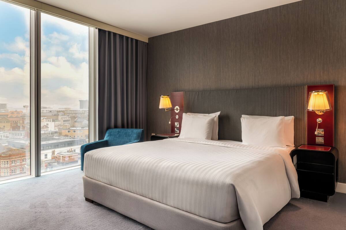Een hotelkamer met een groot bed, twee nachtkastjes, een stoel en uitzicht op de stad door grote ramen.