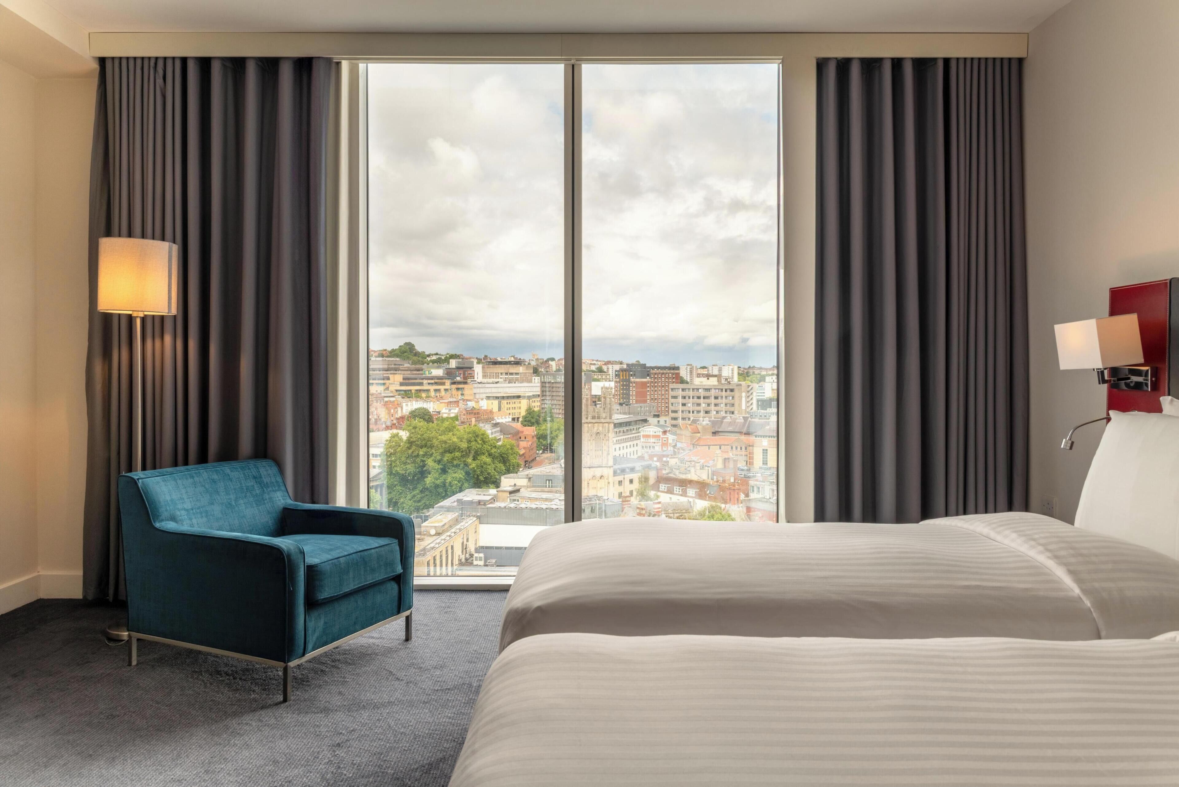 A hotel room with a large window, a bed, a chair, and a lamp.