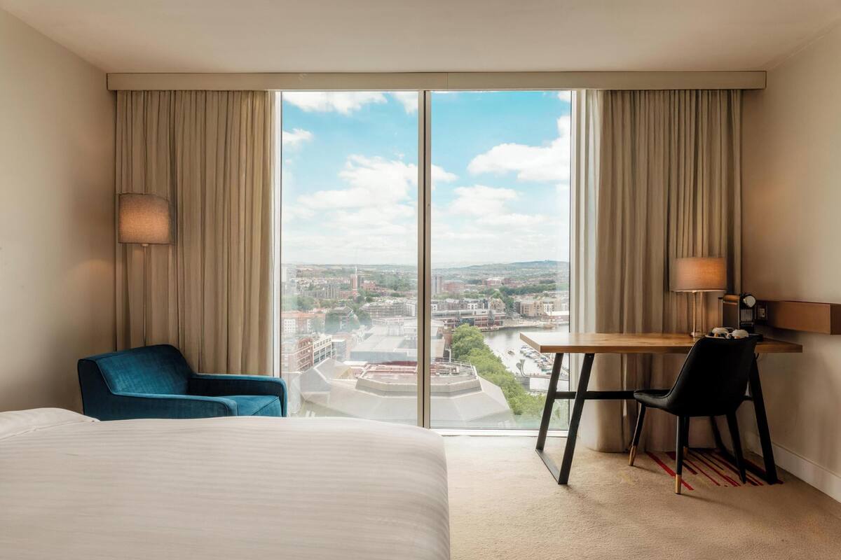 Een hotelkamer met een bed, een bureau, een stoel en uitzicht op een stadslandschap door grote ramen.