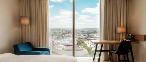 Une chambre d’hôtel équipée d’un lit, d’un bureau, d’une chaise et offrant une vue sur le paysage urbain grâce à de grandes fenêtres.