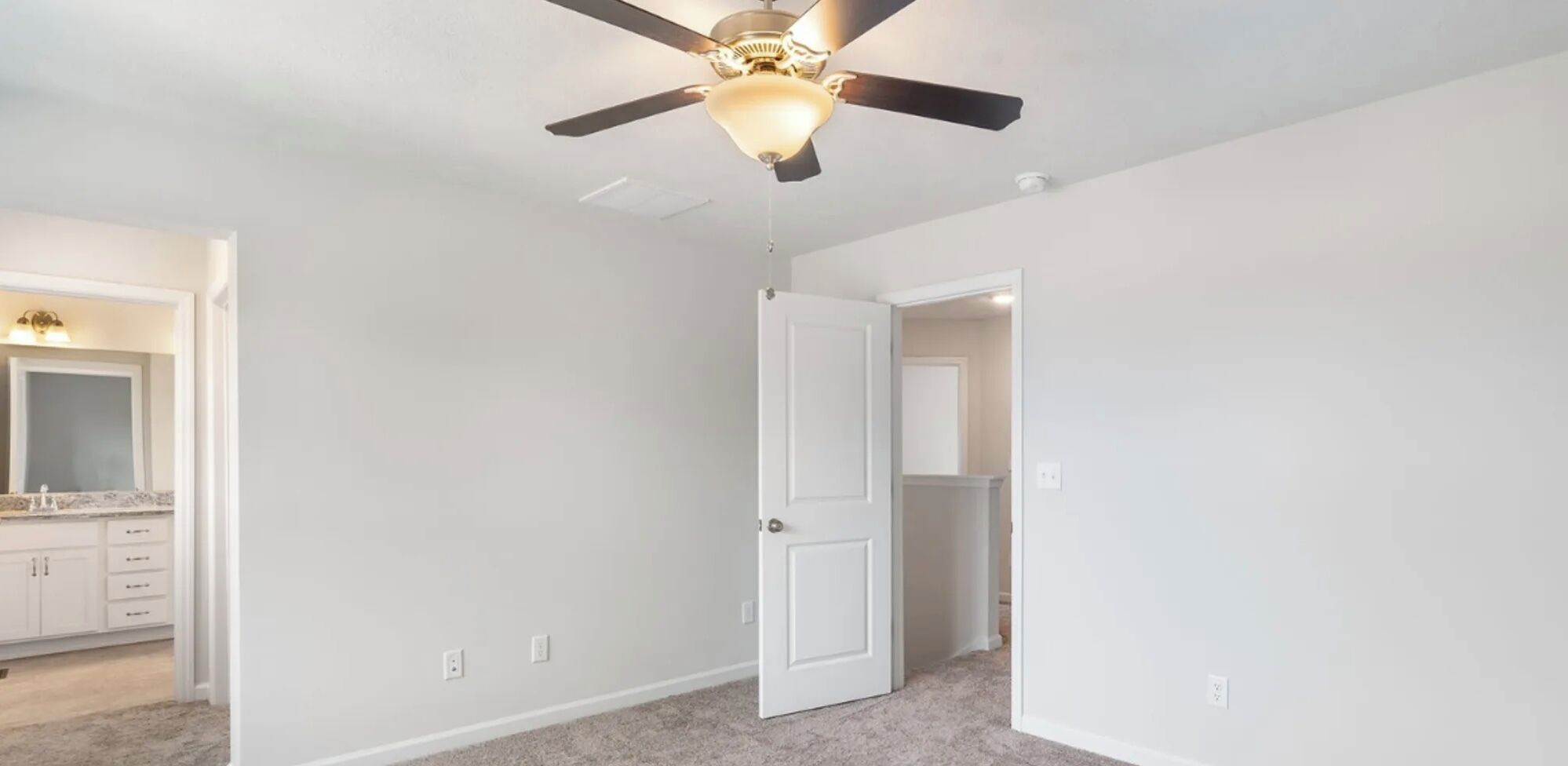 Una stanza con una ventola a soffitto, una porta che conduce in un'altra stanza e una cucina visibile con mobili.