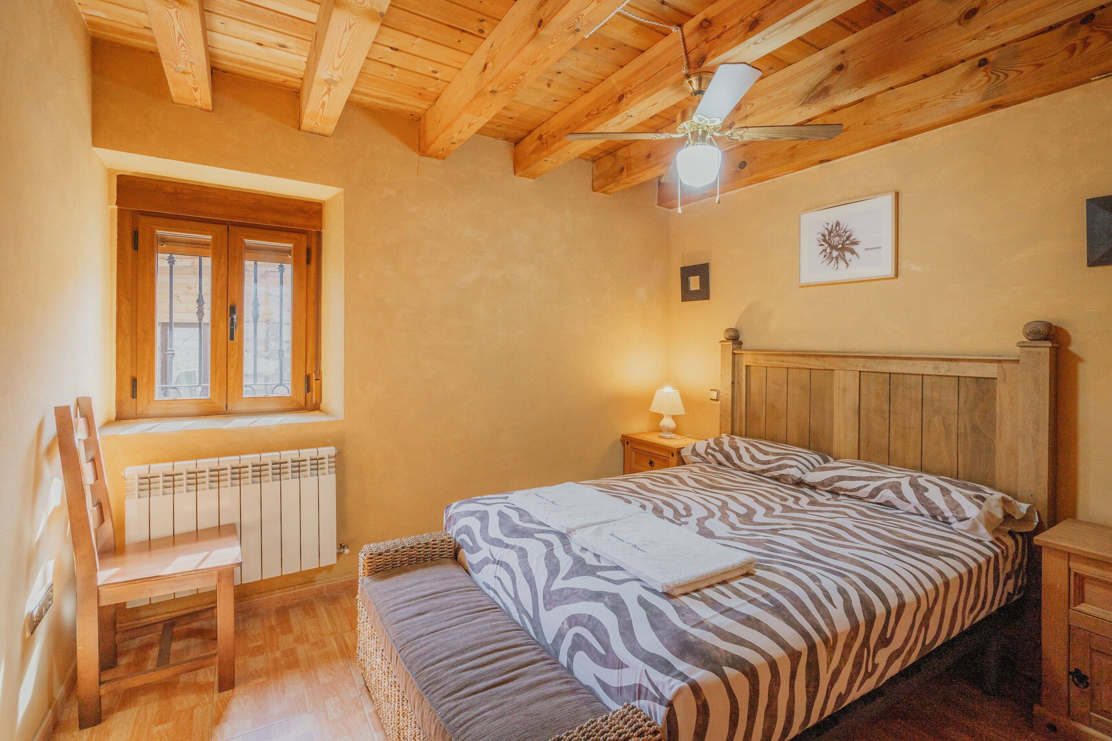 A bedroom with a wooden ceiling, a window with a view, a bed with patterned bedding, a chair, and a bedside table with a lamp.