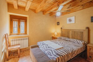 Ein Schlafzimmer mit Holzbalkendecke, einem Fenster mit Blick, einem Bett mit gemusterten Textilien, einem Sessel und einem Nachttisch mit Lampe.