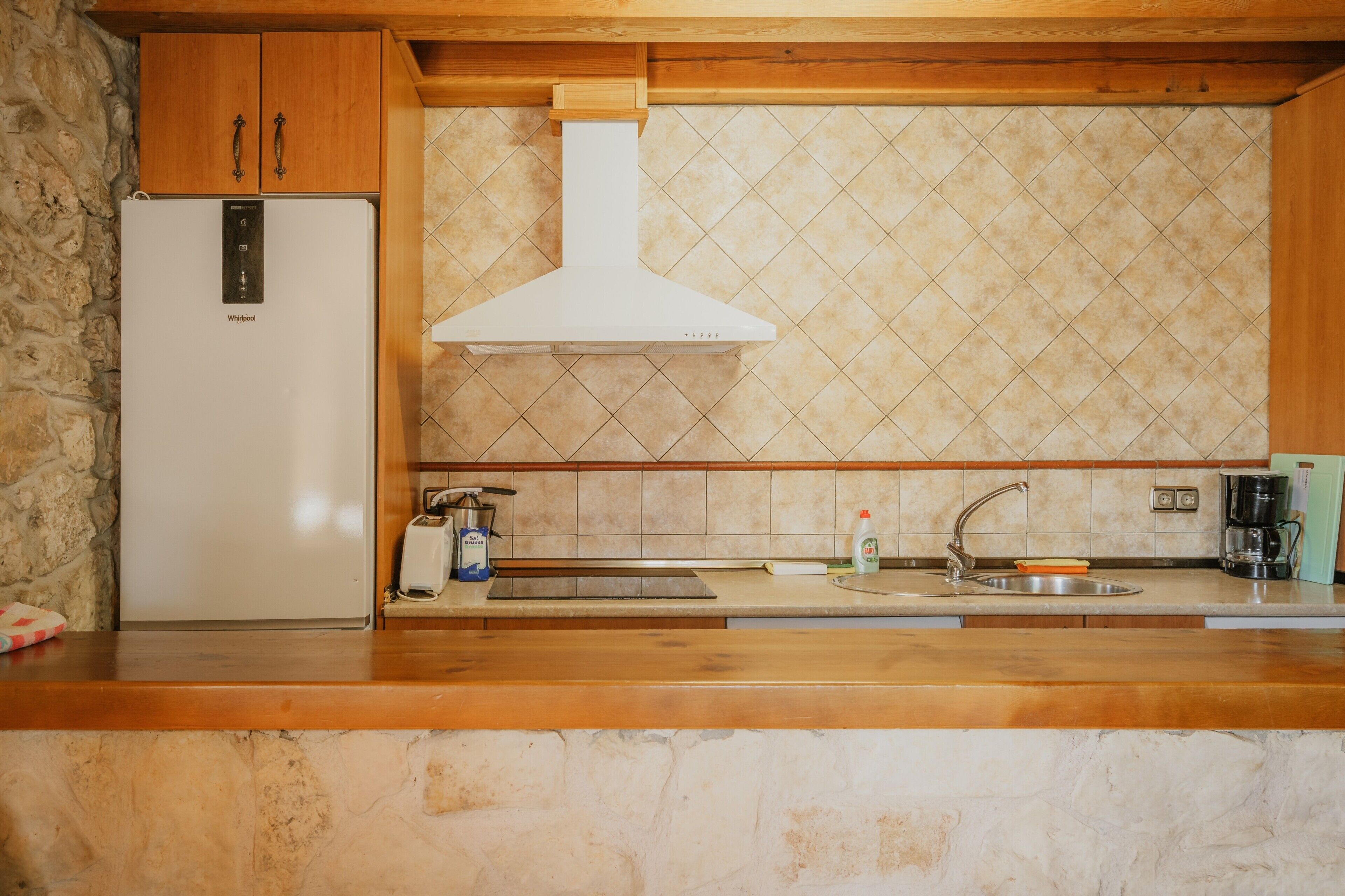 Una cucina con parete in pietra, mobili in legno, frigorifero bianco, lavello in acciaio inossidabile e zoccolo a mosaico.