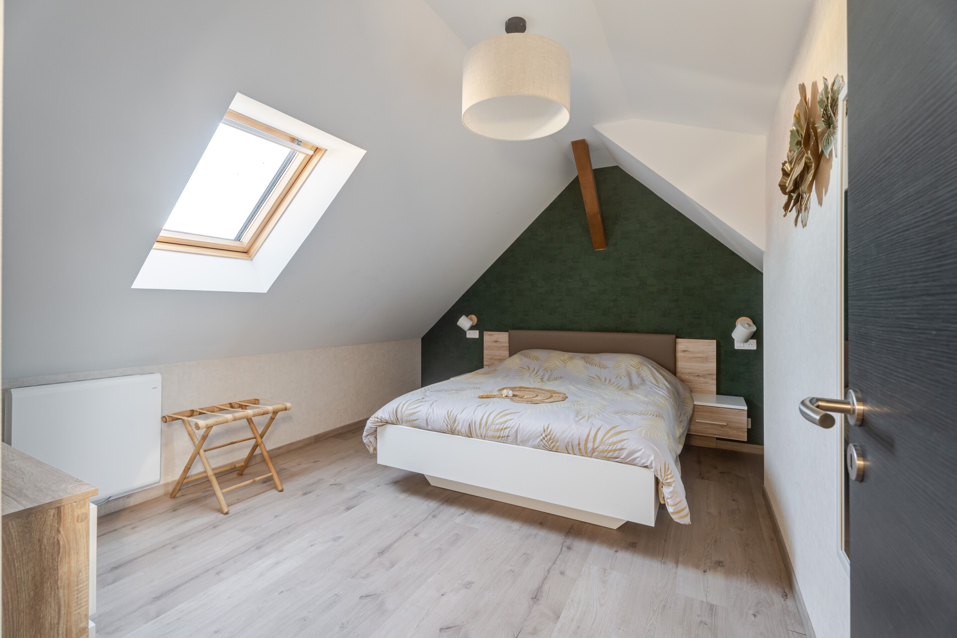 Et soveværelse med skrå vægge, et ovenlys, en seng med et sengetæppe med bladmotiv, et natbord med en lampe og en træskammel.