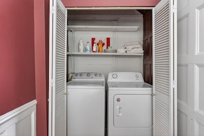 A laundry room with two washing machines, a shelf with laundry supplies, and a door with a red wall. - Front Street Historic Loft | Close to Dining, Bars & Riverwalk (Wilmington)