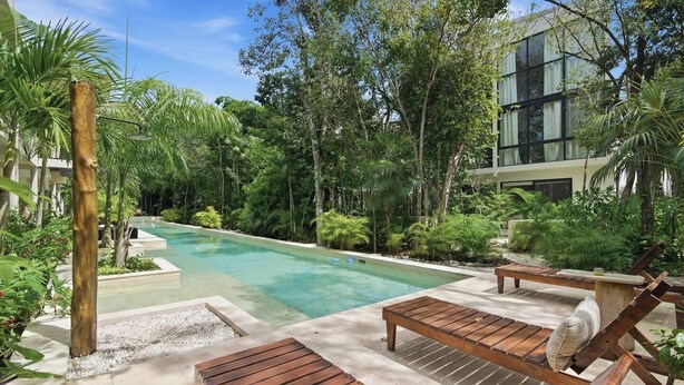 Un espace piscine doté de chaises longues en bois, entouré d’une végétation luxuriante et avec un bâtiment moderne en arrière-plan.