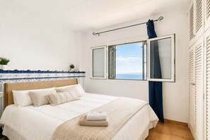Ein Schlafzimmer mit Bett, einem Fenster mit Vorhängen, Blick auf das Meer und einer gefliesten Wand.